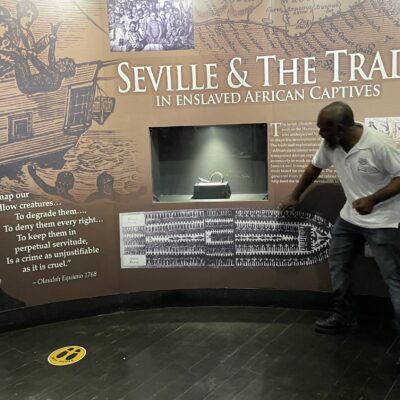 a photo of an exhibition entitled 'Seville and the Trace' with visual imagery of the slave trade. There is a man in front of the exhibition panel pointing to some of the text, which is in white on a brown background