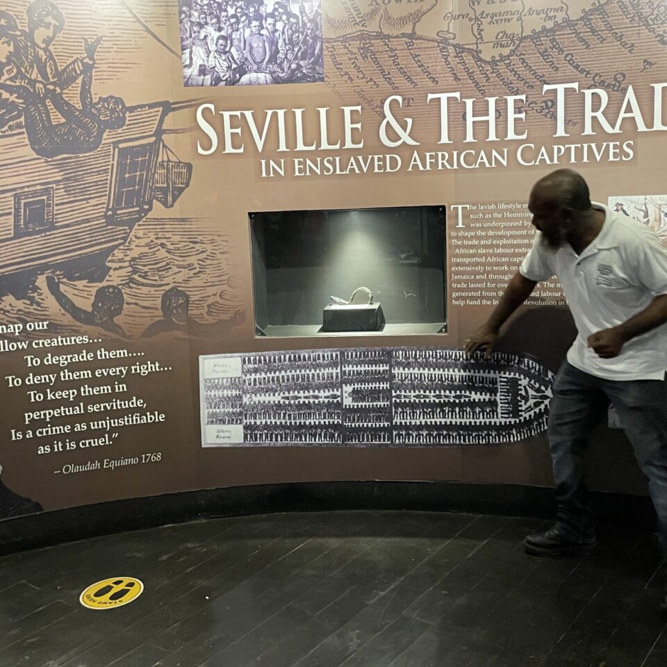 a photo of an exhibition entitled 'Seville and the Trace' with visual imagery of the slave trade. There is a man in front of the exhibition panel pointing to some of the text, which is in white on a brown background