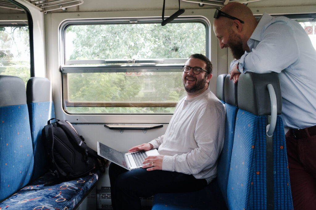 two male passengers on a train, one seated with a laptop, both laughing