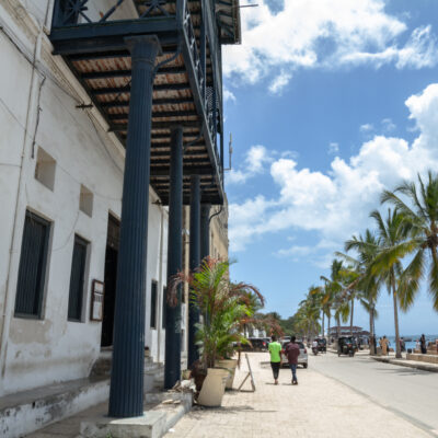 the facade of an old building on the sea front in Stone Town, Zanzibar with palm trees along a path and blue columns on the front of the building