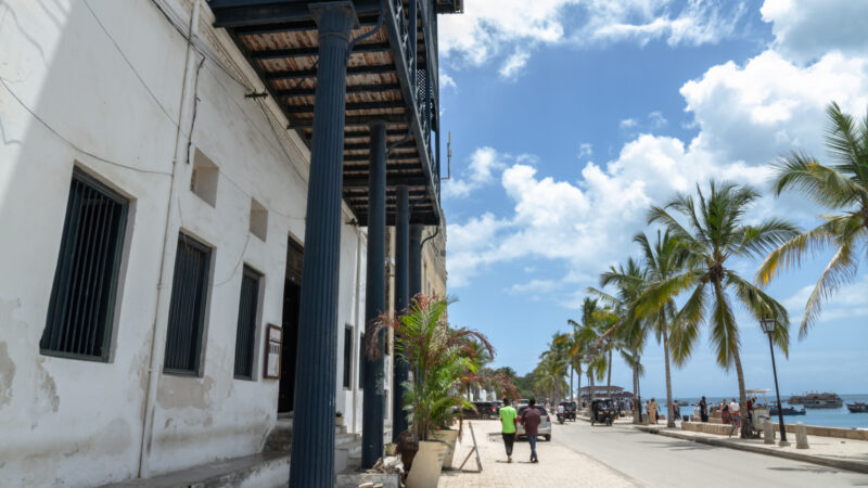 the facade of an old building on the sea front in Stone Town, Zanzibar with palm trees along a path and blue columns on the front of the building