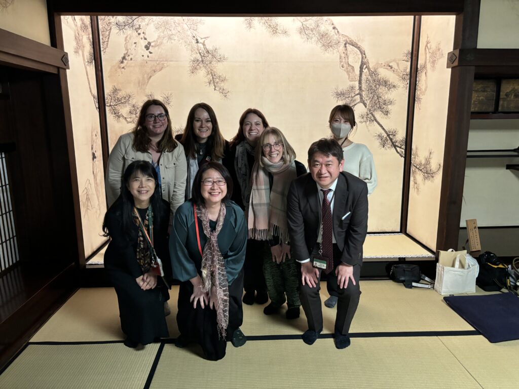 a group of people smiling at the camera in a Japanese room with a screen decorated with Japanese foliage. One of the group is part of TAP INTO grant awarded