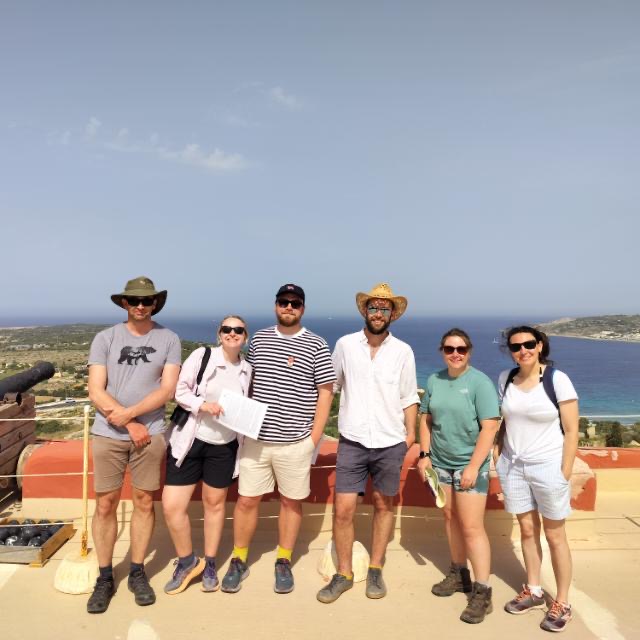 A group of people smiling at the camera, with the backdrop of blue sea and bright blue sky, taken in Malta