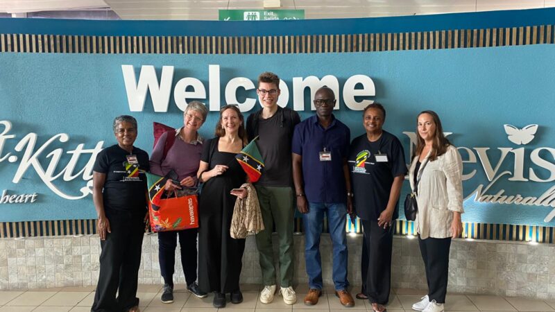a group of people standing in front of a sign saying welcome to st kitts and nevis, smiling at the camera