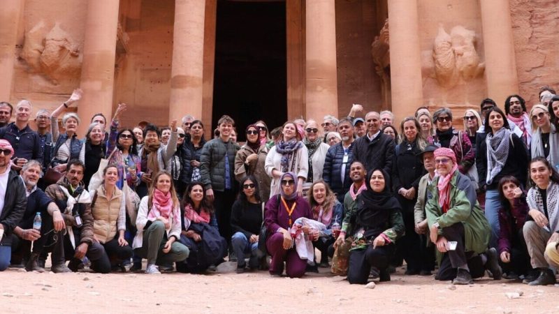 a large group of people smiling at the camera in front of The Treasury, a large temple carved from red stone in Petra, Jordan