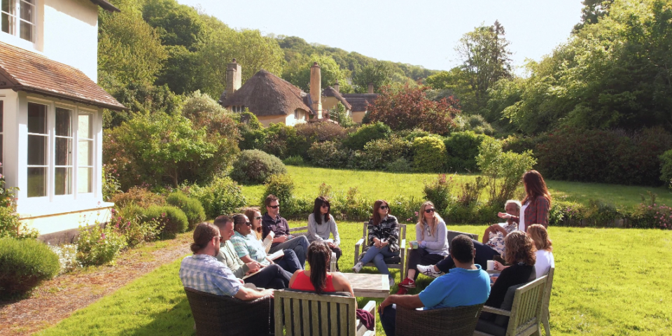 A large group of people sitting in a circle outside a cottage in the sunshine