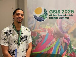Nigel youth delegate to GSIS he is wearing a traditional headpiece and face adornment and a black and white shirt he is standing in front of a brightly coloured poster for the GSIS summit 2025