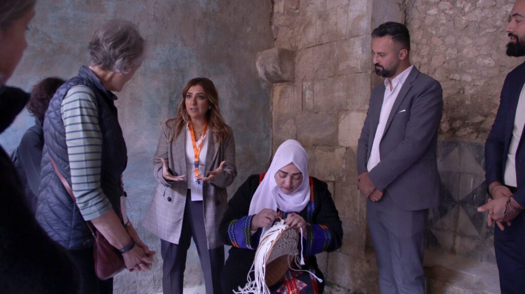 a woman in a white hijab is seated making tassels by hand. a woman is standing next to her explaining her work to another standing visitor. a man also stands to the other side of the seated woman. they are in a stone room in As-Salt Jordan