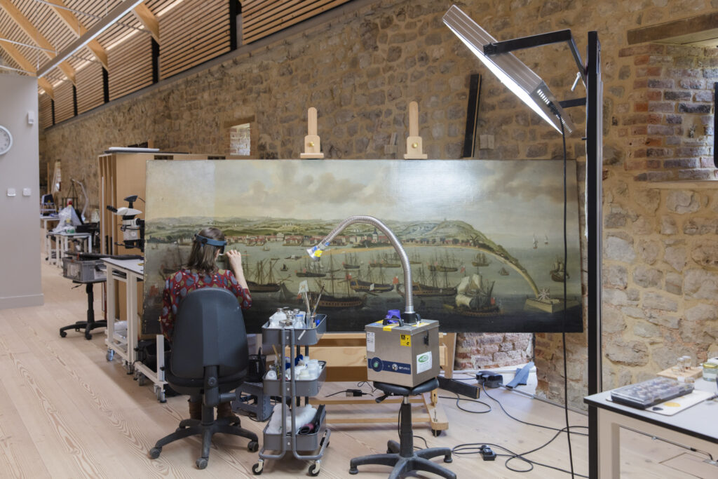 the interior of a painting conservation studio with a large canvas on an easel undergoing cleaning, with one female conservator seated with her back to the camera. Alongside her is a trolley of technical conservation tools.
