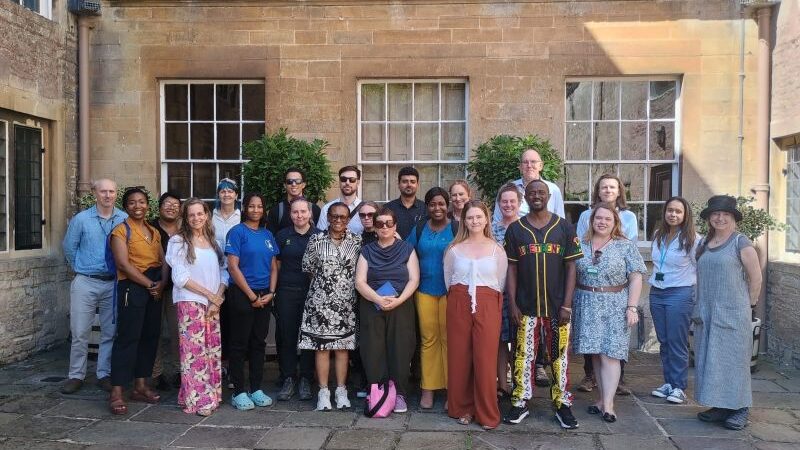 a group of people in a formal pose smiling at the camera. They are standing in front of 3 large windows which are part of an historic house at Dyrham Park and meeting for the INTO RISE programme