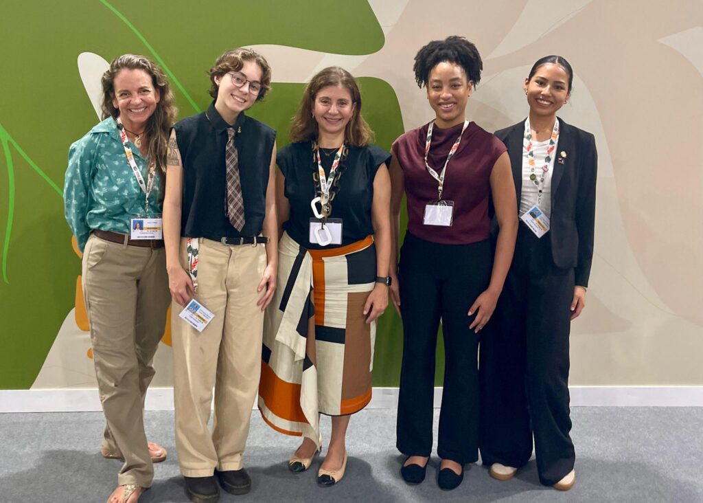 five women standing and smiling at the camera, they are in front of a wall with a huge leaf motif at COP30 in Brasil
