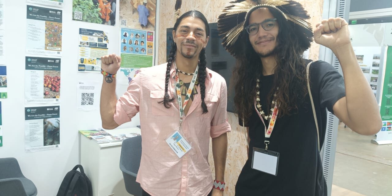 two young men with indigenous headpieces raising their fists to the camera in a gesture of solidarity at the INTO booth at COP30