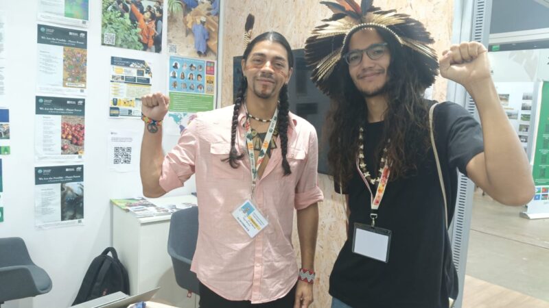 two young men with indigenous headpieces raising their fists to the camera in a gesture of solidarity at the INTO booth at COP30