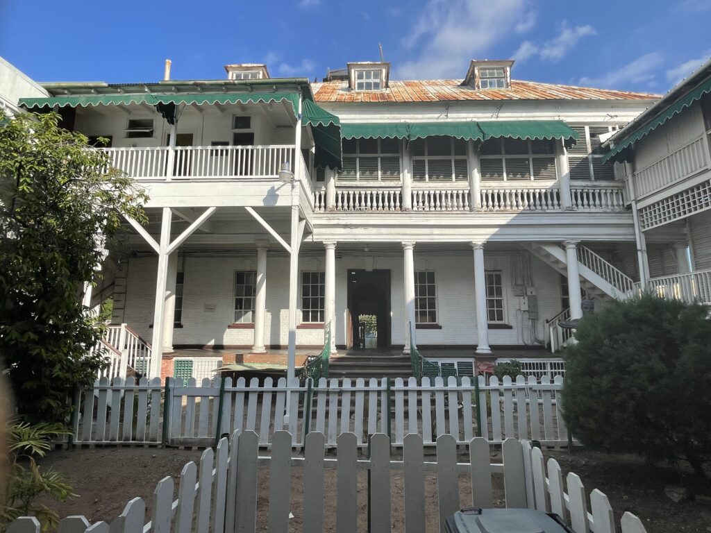 the exterior of Headquarters House in Kingston Jamaica. It is a white wooden house with a first floor veranda and green sun shades about the veranda. There is a white picket fence in front of the building.