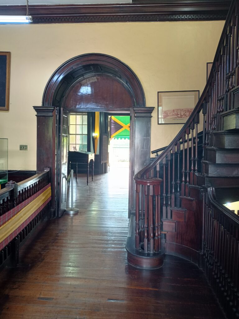 The interior of Headquarters House in Kingston Jamaica. It has dark ploished wood floors, an arched doorway and the foot of a curved wooden staircase is on the right of the photo