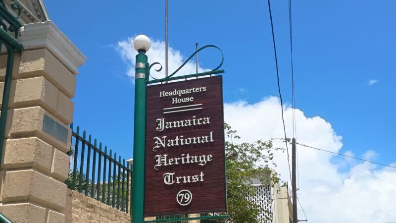 a brown and white sign reading 'Jamaica National Heritage Trust' on a green pole with blue skies