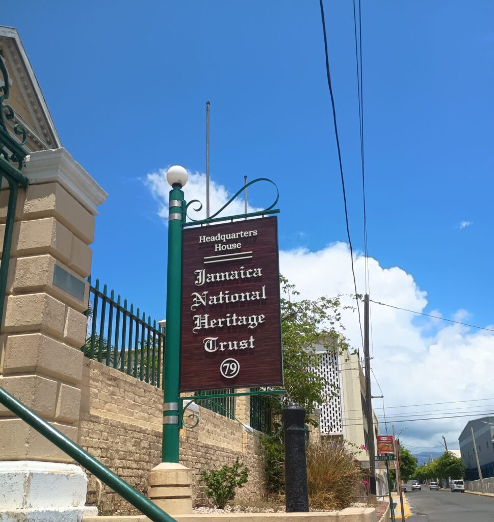 a brown and white sign reading 'Jamaica National Heritage Trust' on a green pole with blue skies