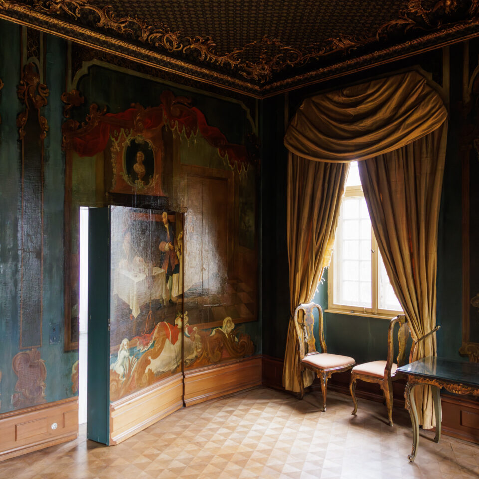 a dark room in an historic building with a patterned wooden floor and dramatic drapes at the window