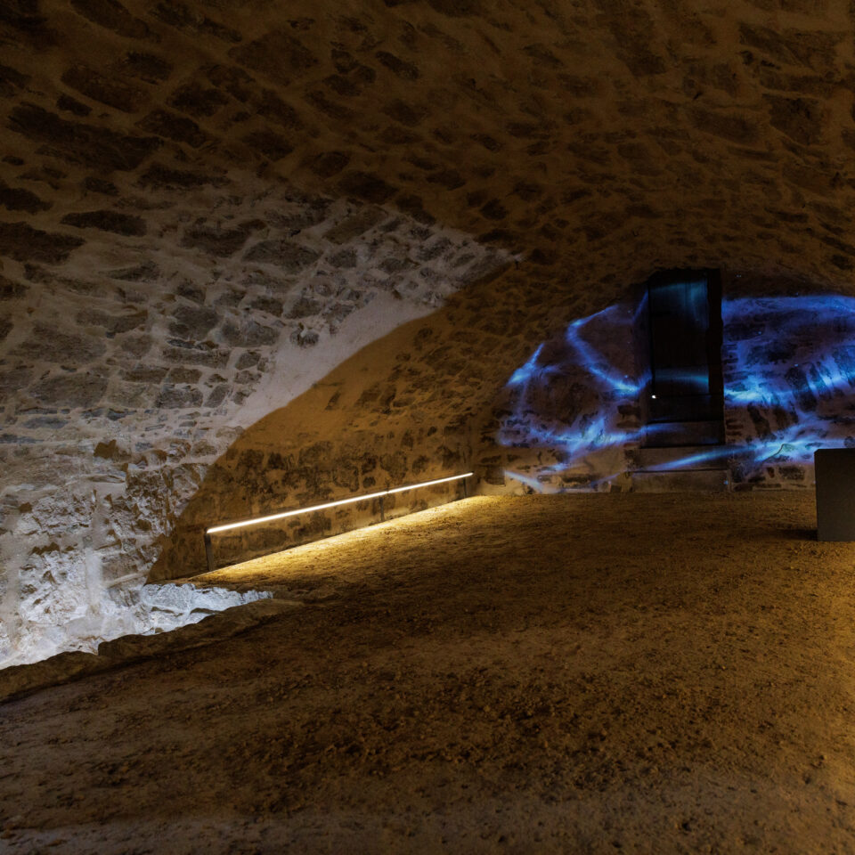 a dark stone interior with a curved ceiling like a cave, with a projection on the end wall in blue giving the impression of water and ripples