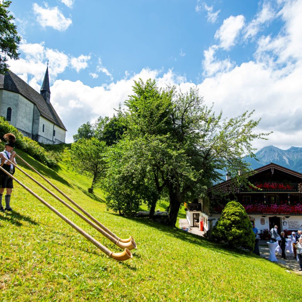 a view of the Bavarian alps with a traditional alpine inn, a white church and some people in traditional Bavarian dress playing long horn-shaped musical instruments