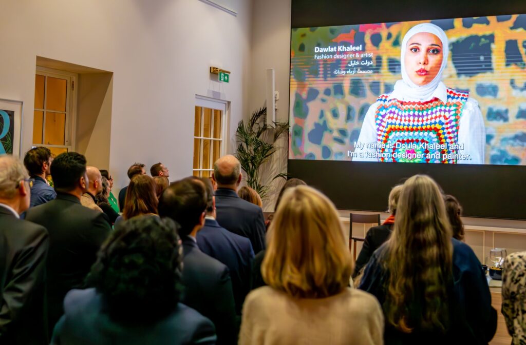 A group of people watching a large video screen. There is a woman talking on the screen, wearing a hijab