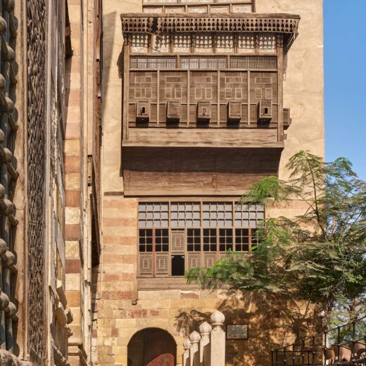 The facade of a stone building in Cairo, Egypt with elaborate wooden features. There is a small tree in the foreground