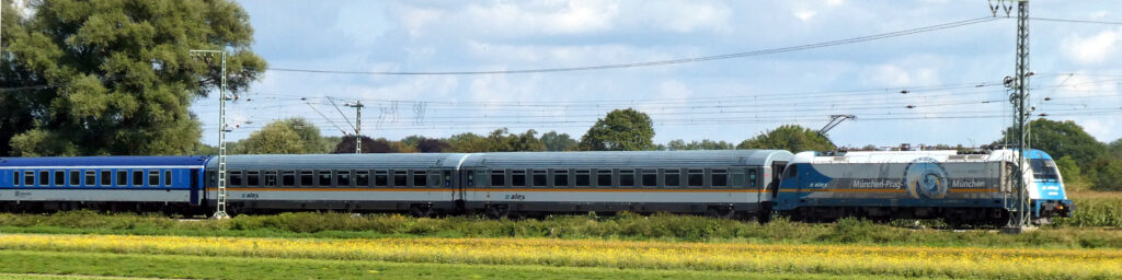 a German train with an engine and several carriages. There is blue sky above the train and a field with yellow flowers in the foreground