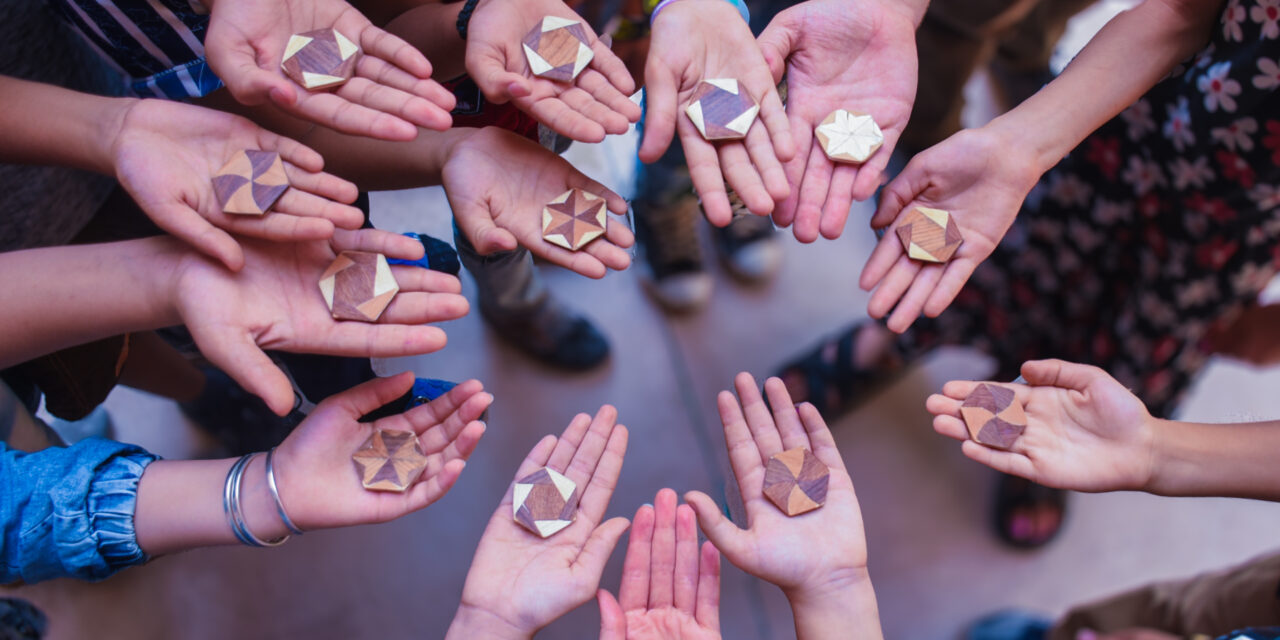 a circle of upturned children's hands, each holding a geometric wooden tile with Arabic designs