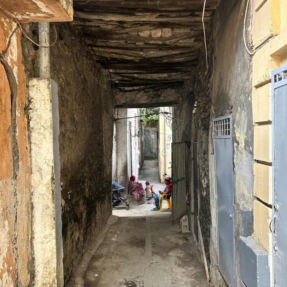 View of women sitting in the narrow streets of Mutsamudu through a tunnel supported by mangrove poles and coral stone.