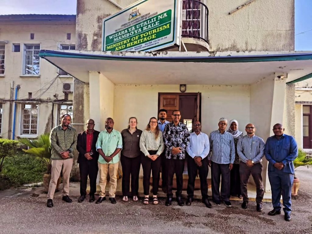 a group of people standing in front of a cream-coloured building with a green sign that reads 'ministry of tourism and heritage' in Stone Town Zanzibar